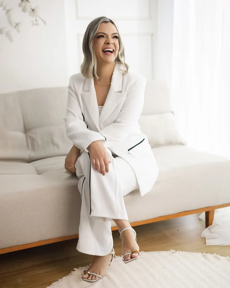 Dra. Camila Ferreira, de terno branco, sentada num sofá claro e sorrindo